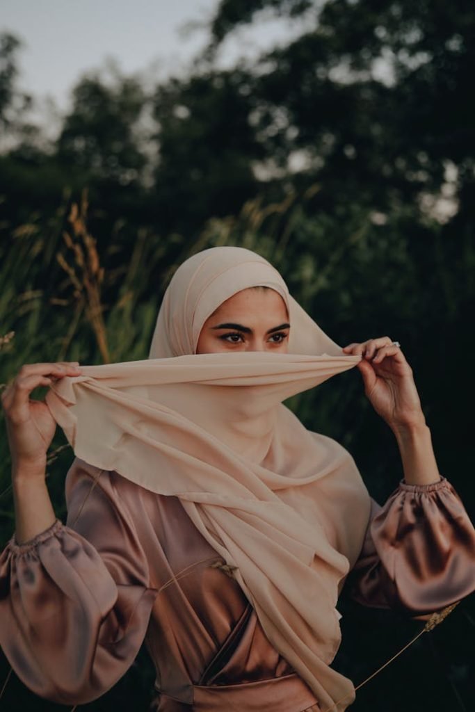 A woman in a silk hijab holding it gently, captured outdoors during evening light.