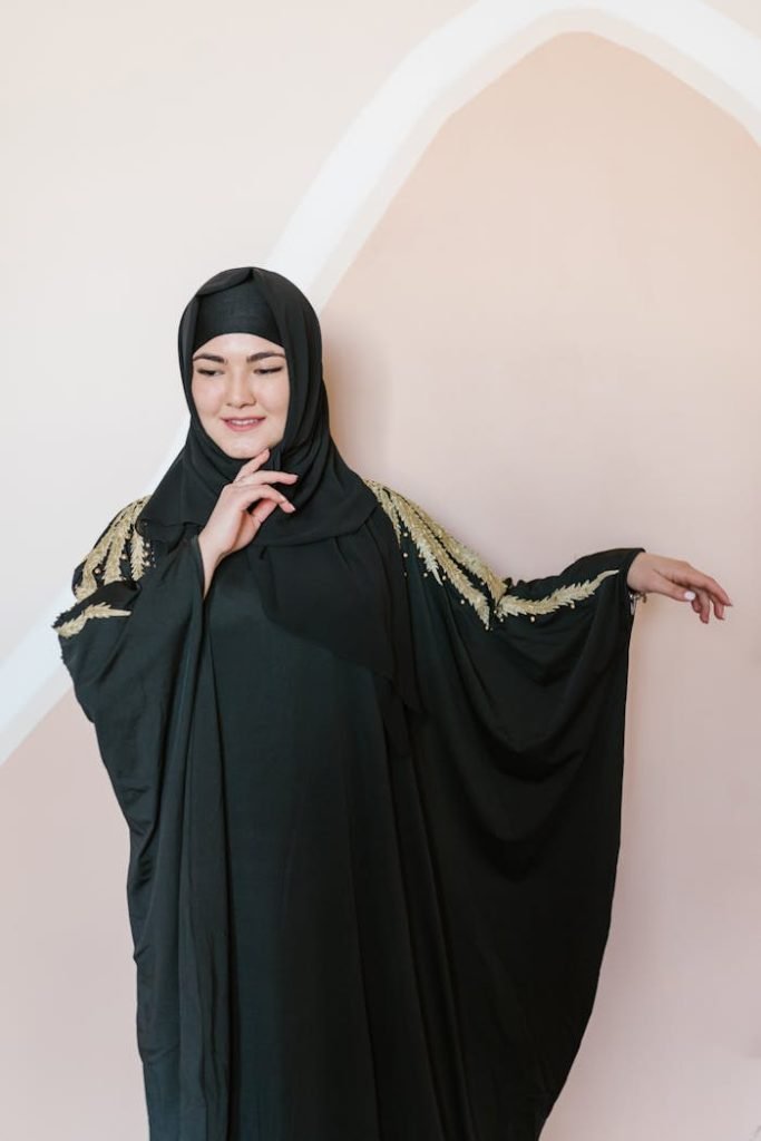 Graceful woman in a black abaya with gold embroidery poses elegantly indoors.